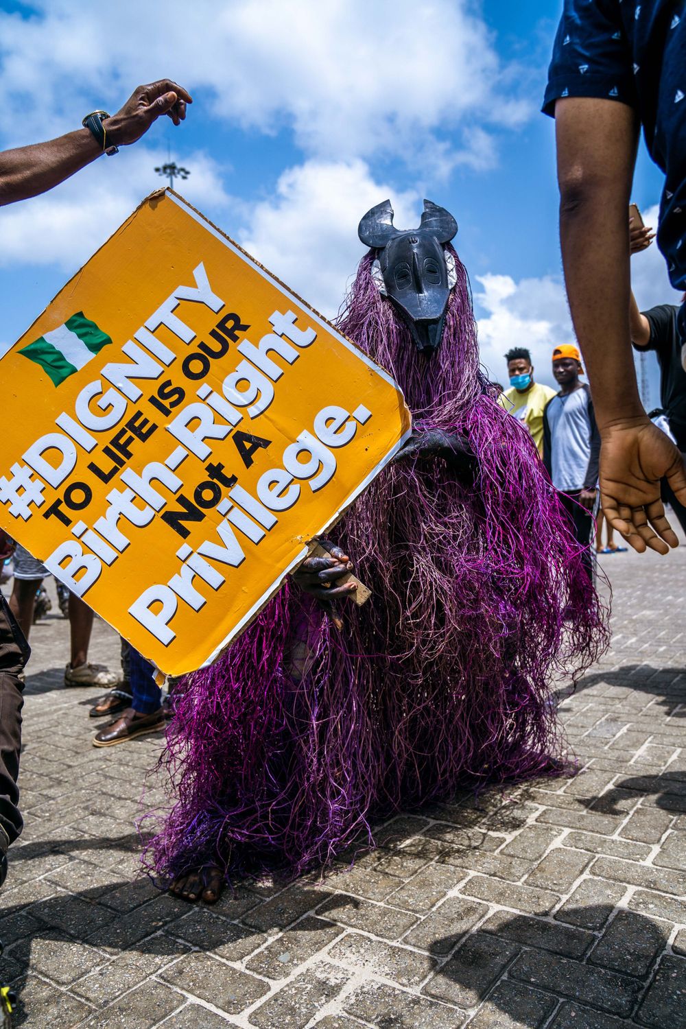 Purple-costumed demonstrator with horned mask holding yellow protest sign on brick pavement.