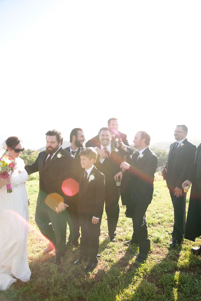 Wedding party gathered outdoors in formal attire laughing together in bright sunlight.