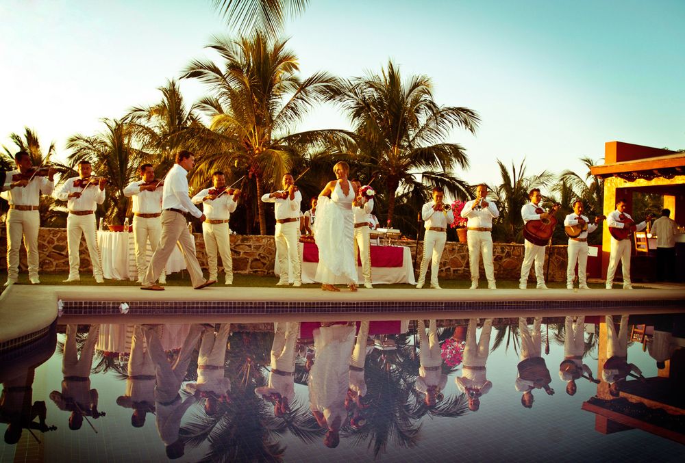 Wedding guests line up along a pool's edge at sunset with palm trees and reflections in the water.