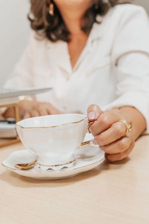 Elegant white teacup with gold trim held by hands wearing rings on a wooden table.