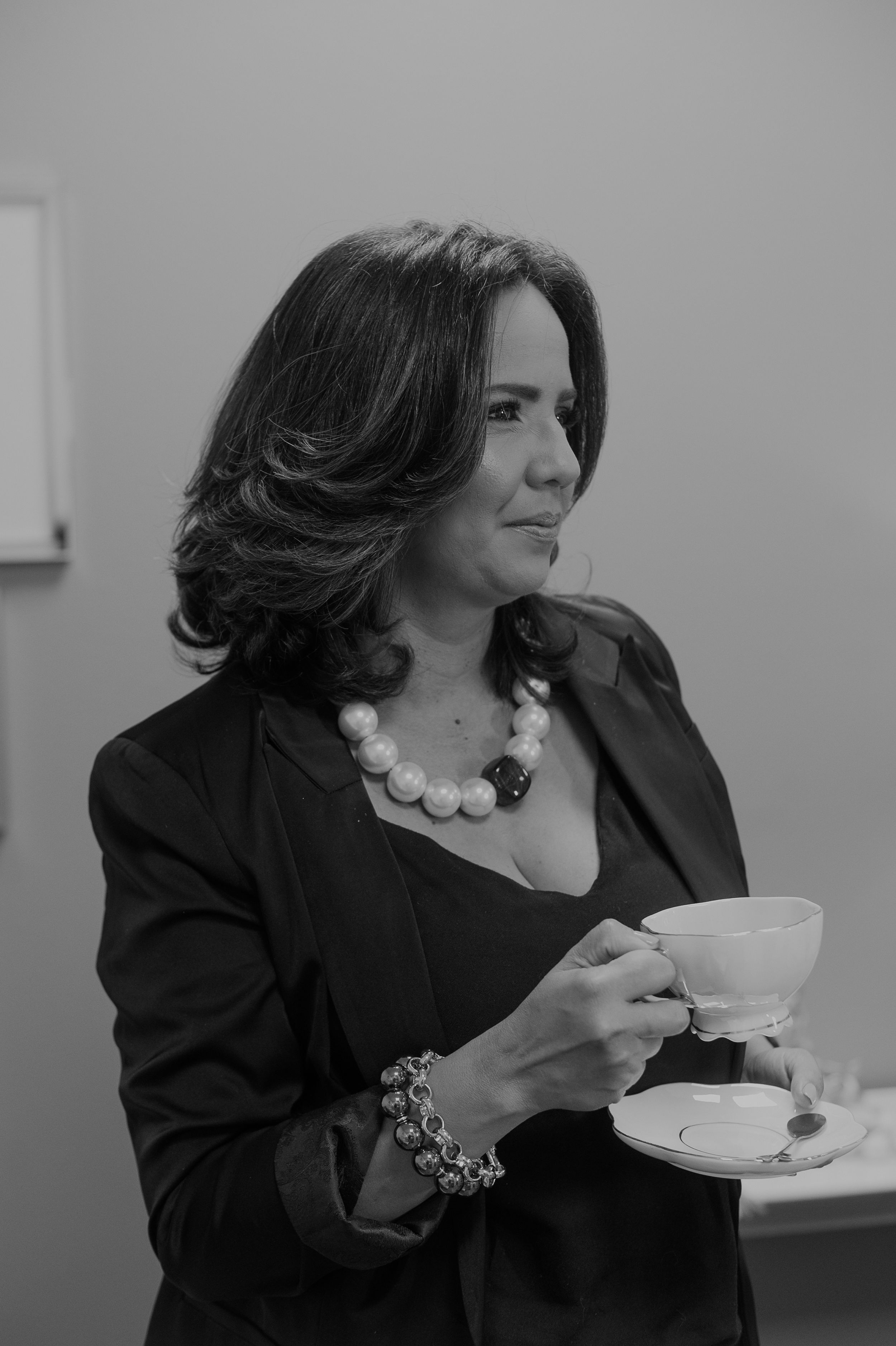 Professional woman in black blazer and pearl necklace holding teacup in elegant black and white portrait.