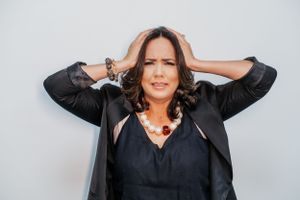 Expressive portrait of woman in black jacket and pearl necklace with hands behind head.