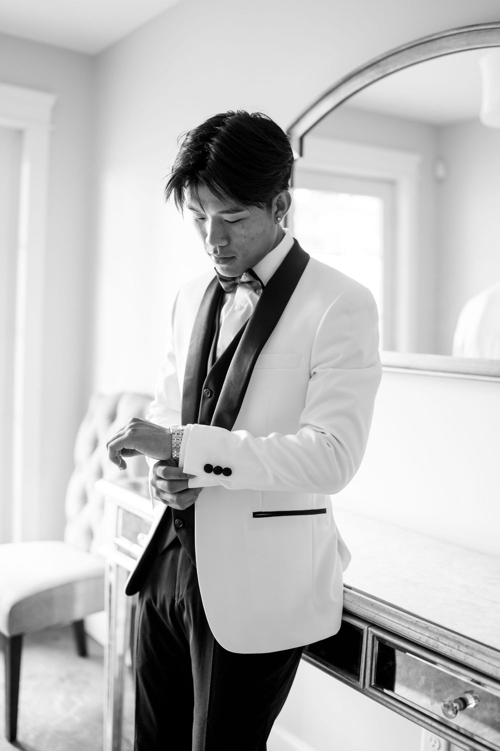 Black and white photo of groom in white tuxedo adjusting cufflinks before wedding ceremony.
