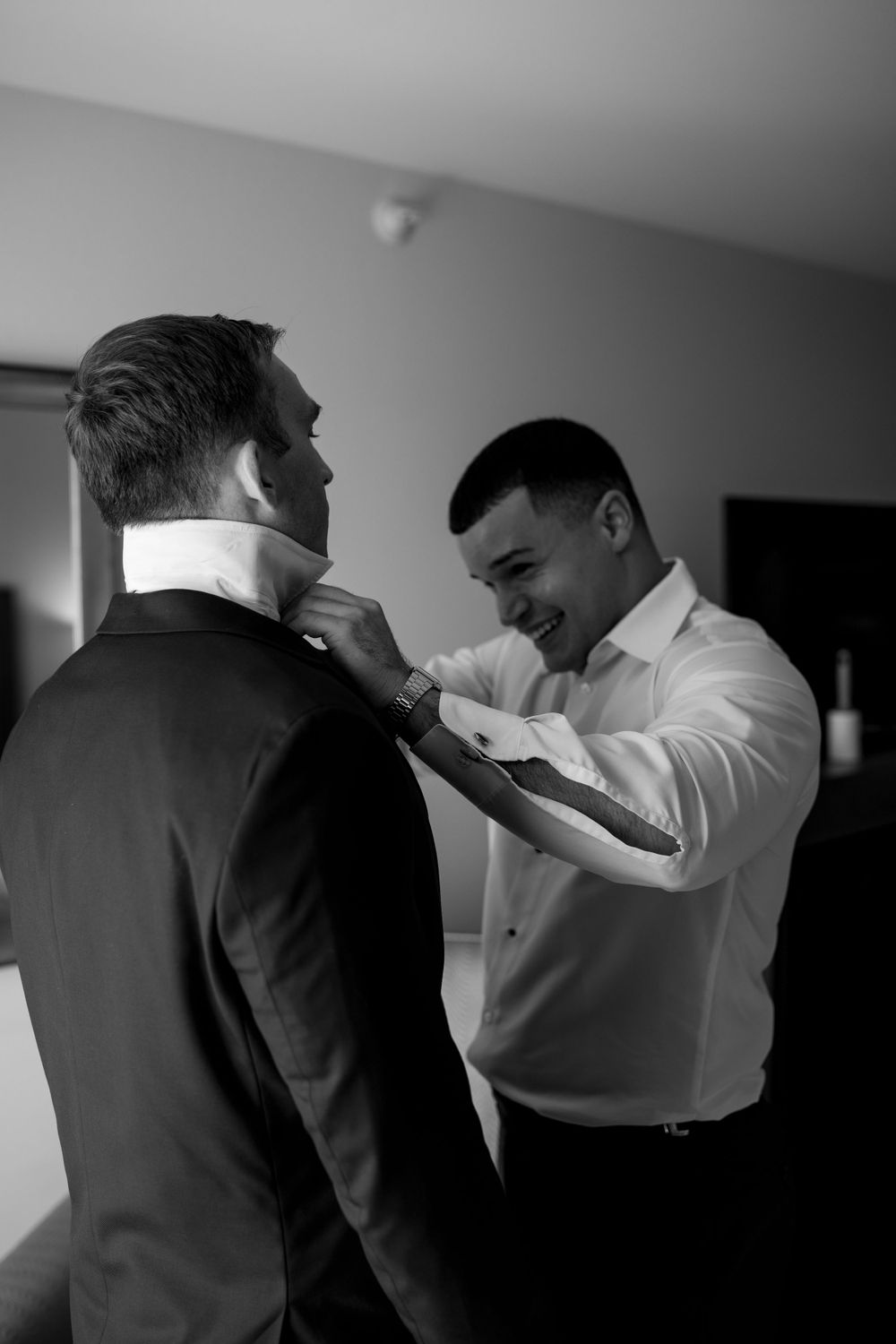 Groomsman helps groom adjust collar and tie while smiling during wedding day preparations.