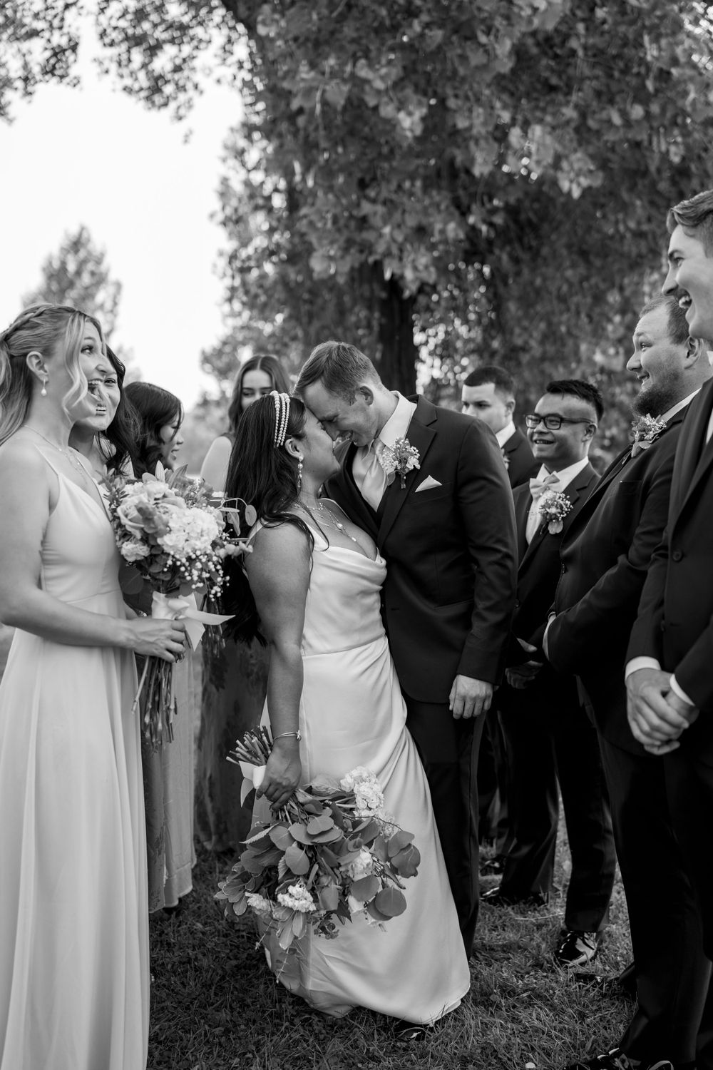 Bride and groom share a kiss surrounded by smiling wedding party in black and white photo.