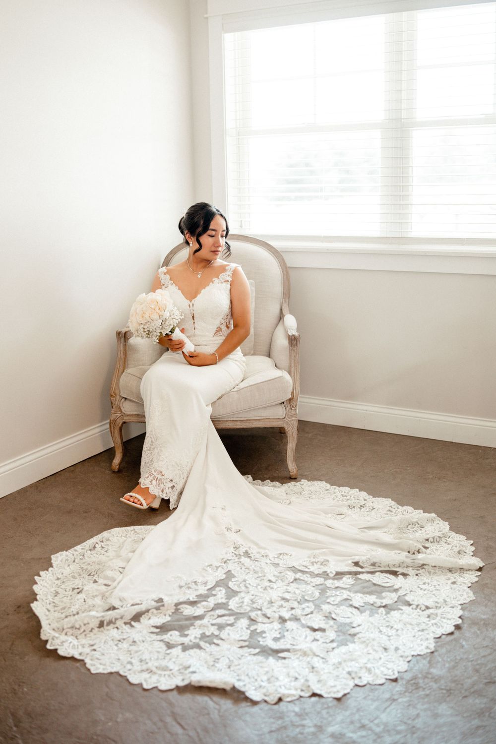 Elegant bride in fitted lace wedding gown with long train sits in vintage chair by window.