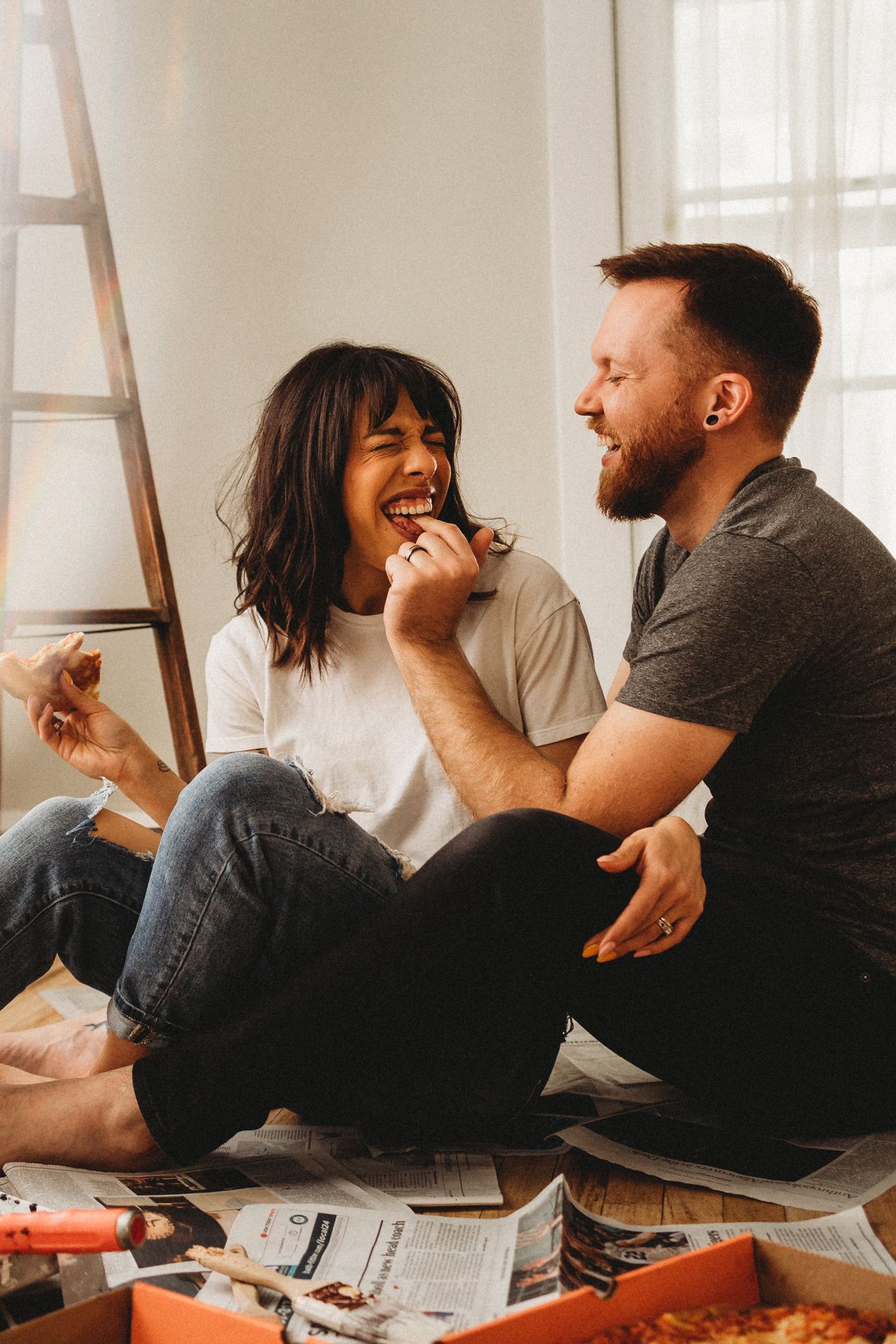 Couple laughing while sharing pizza on floor of bright room.