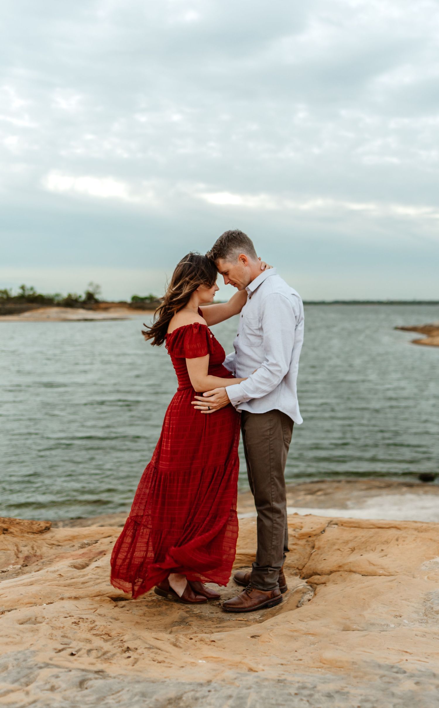 A romantic sunset beach photoshoot with a flowing red dress against a cloudy waterfront backdrop.