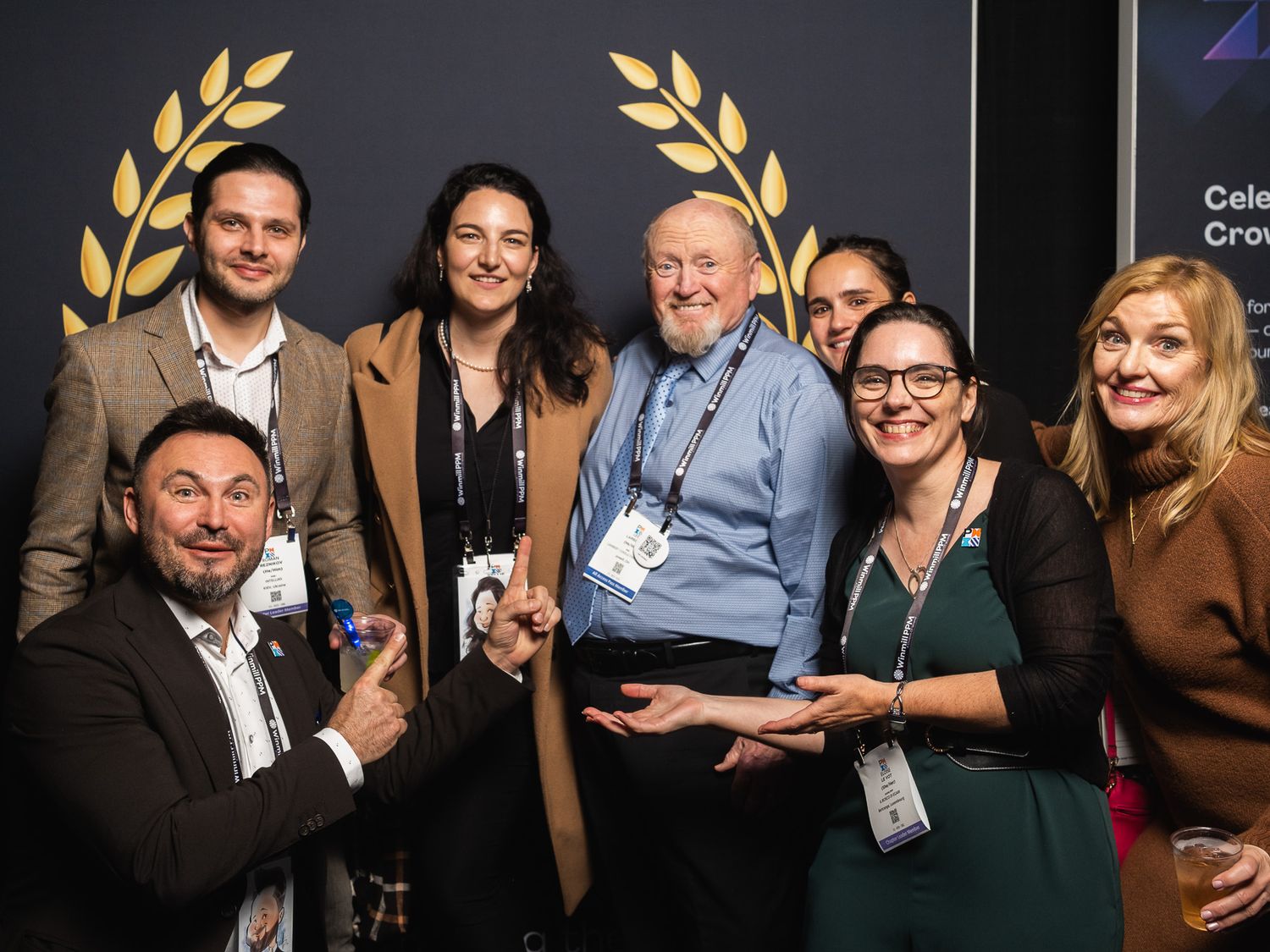 A group of professionals pose together at a business event with golden laurel wreath decorations on the wall behind them.