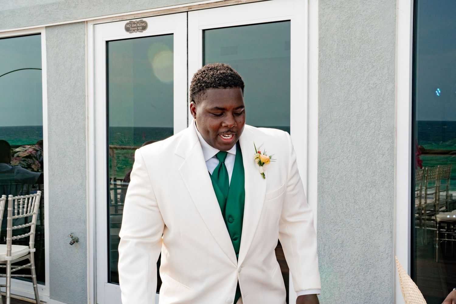 A person in a crisp white suit jacket with an emerald green tie and boutonniere smiles outside glass doors.
