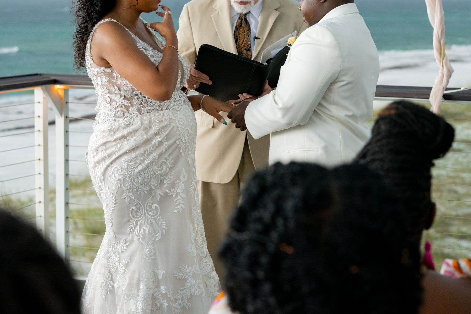 A couple exchanges vows during a beachside wedding ceremony with a white lace bridal gown in view.