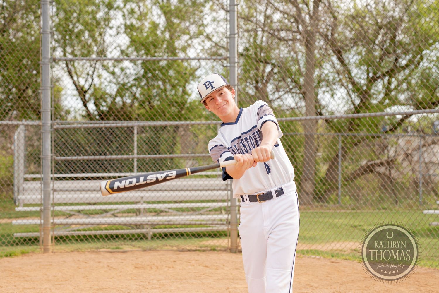 Baseball player in white uniform swings bat during practice with chain link fence behind.