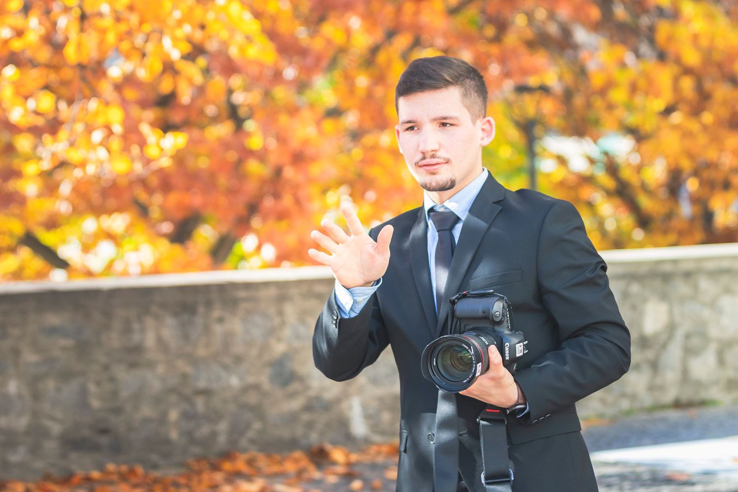 Professional photographer in suit holding camera with autumn foliage background.