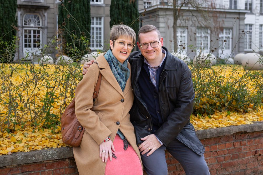 A couple sits on a brick wall with fall foliage and a stately building behind them.