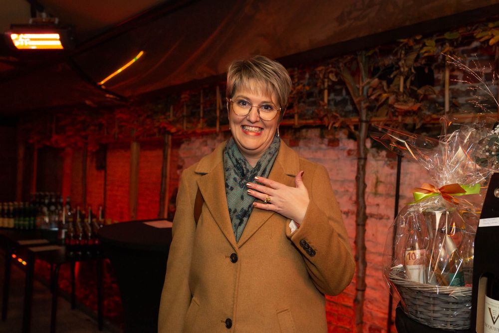 A person in a camel coat smiles while holding a celebratory gift in a warm-lit indoor setting.