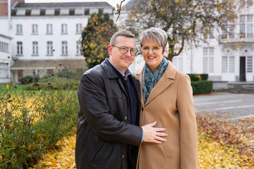 A couple embraces outdoors in autumn with yellow foliage and a white building in the background.