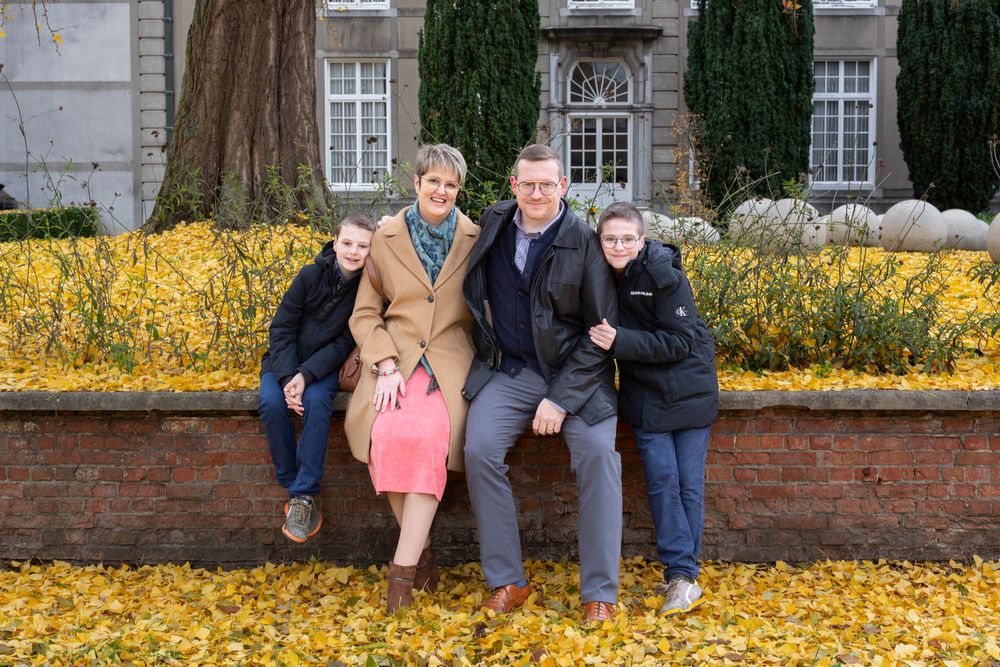 A family group sitting on a brick wall surrounded by yellow autumn leaves and cypress trees.