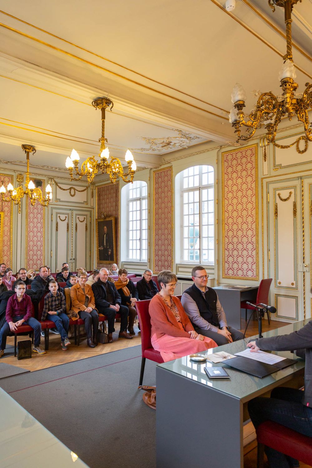 An ornate historic hall with golden chandeliers and pink wallpaper filled with seated attendees.