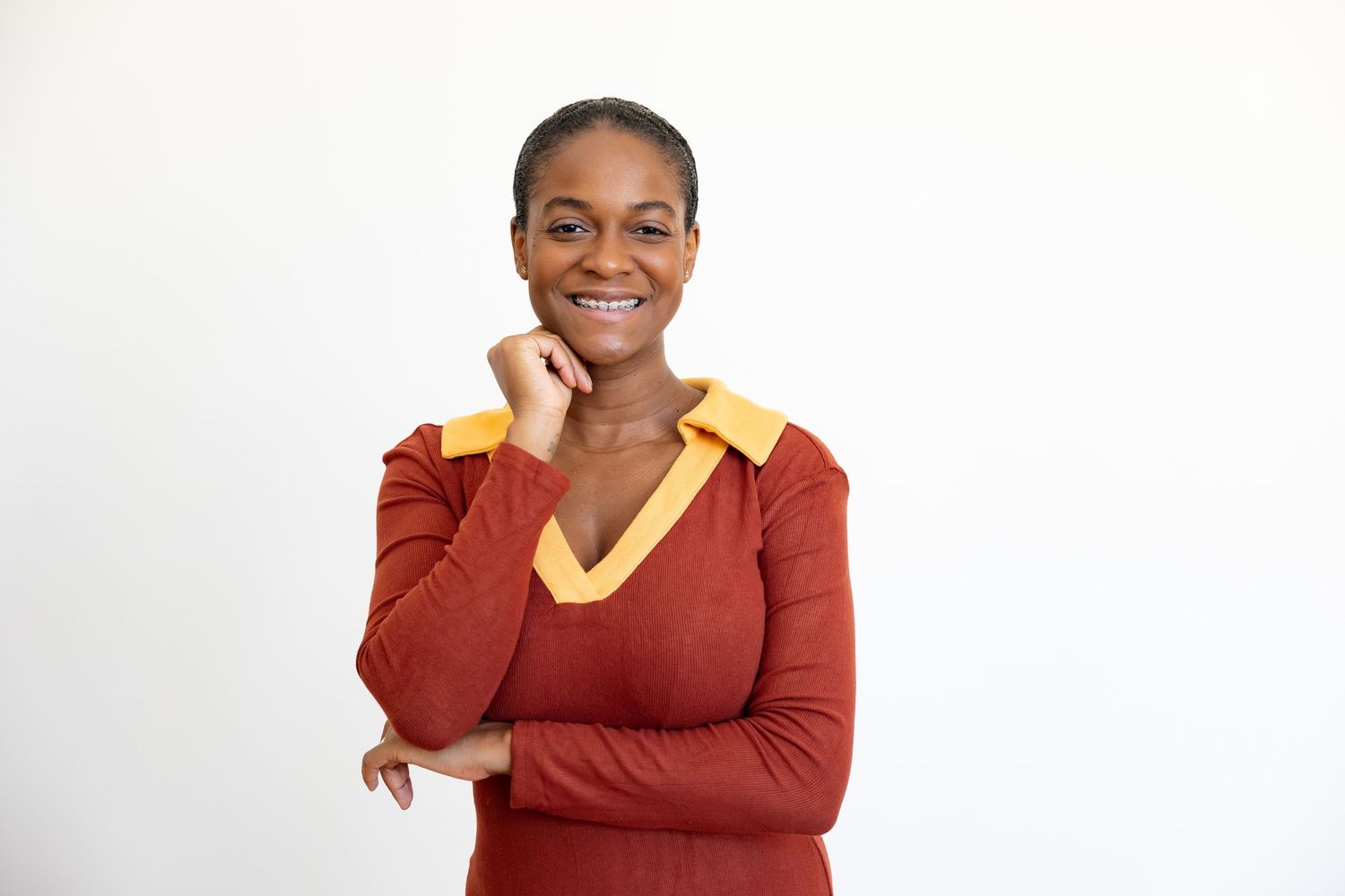A smiling Black woman in a red and gold v-neck top poses thoughtfully with hand near chin on white background.