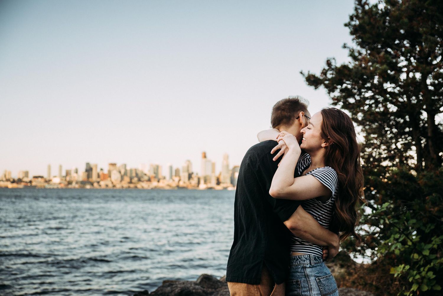Couple snuggles for portraits with Seattle skyline behind them.