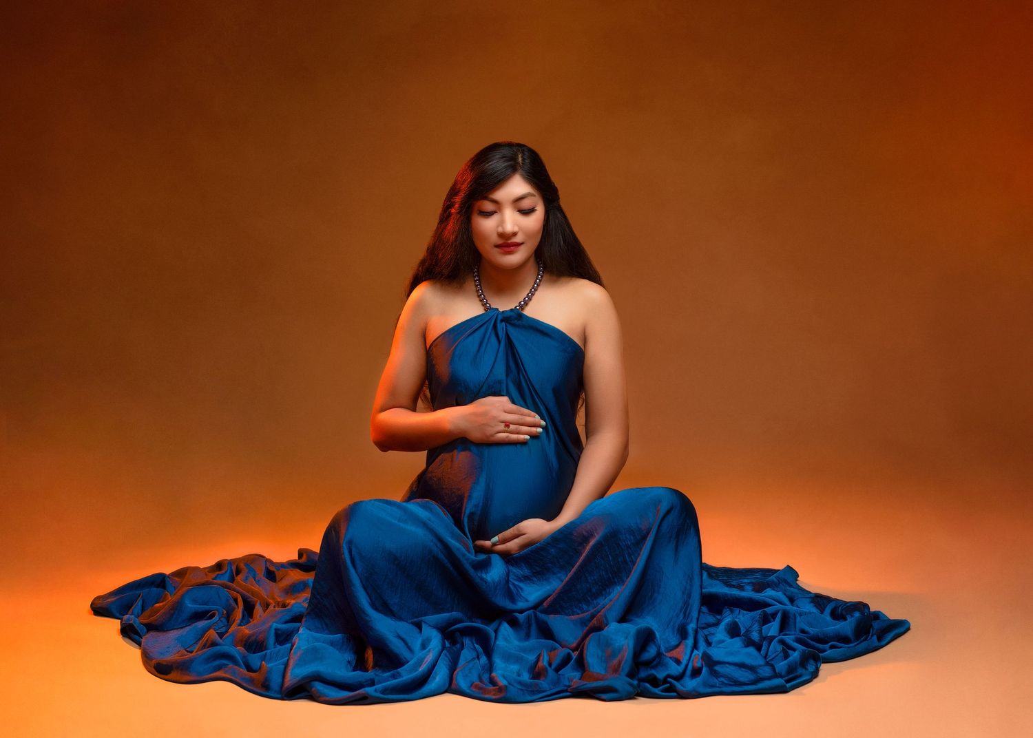an expecting mother sits criss cross in a flowing blue dress with her hands on her belly