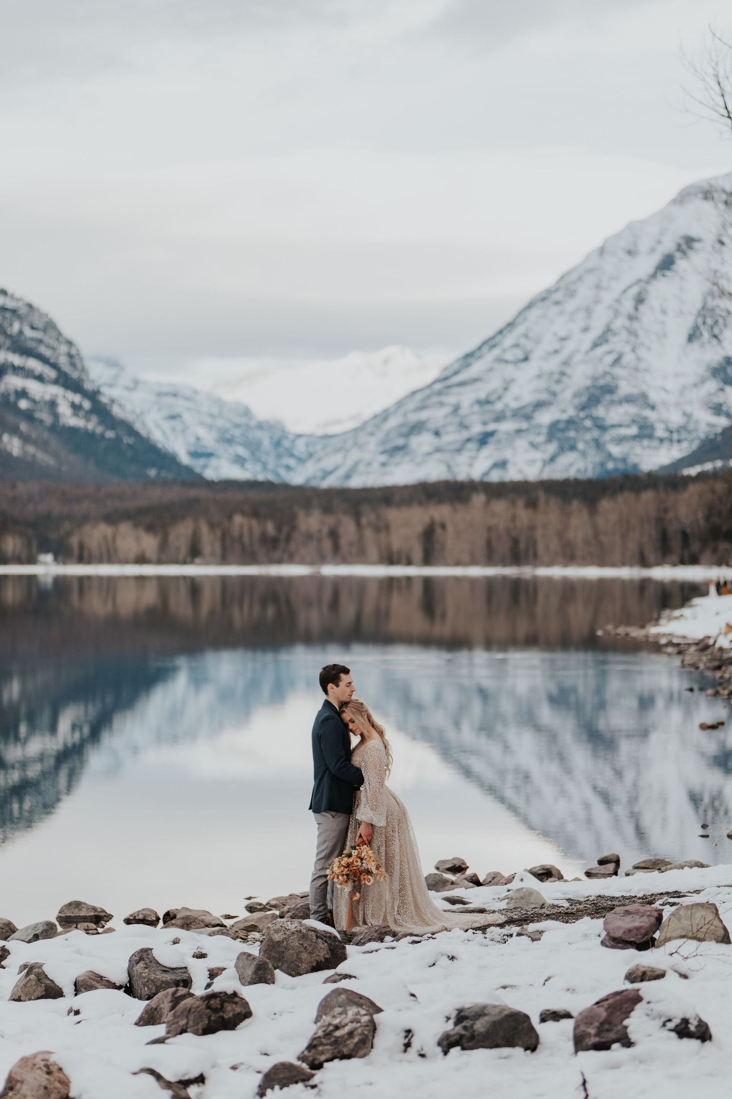 Eloping couple in glacier national park embracing on the edge of a reflective lake and snow covered mountains