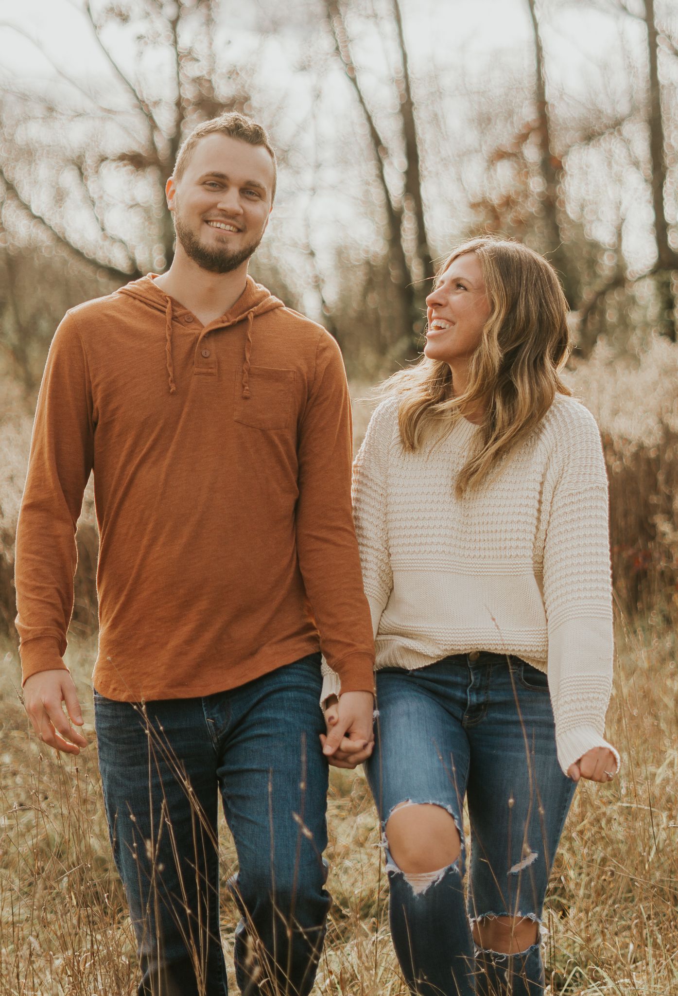 Couple walking hand in hand through tall grass in autumn clothing.