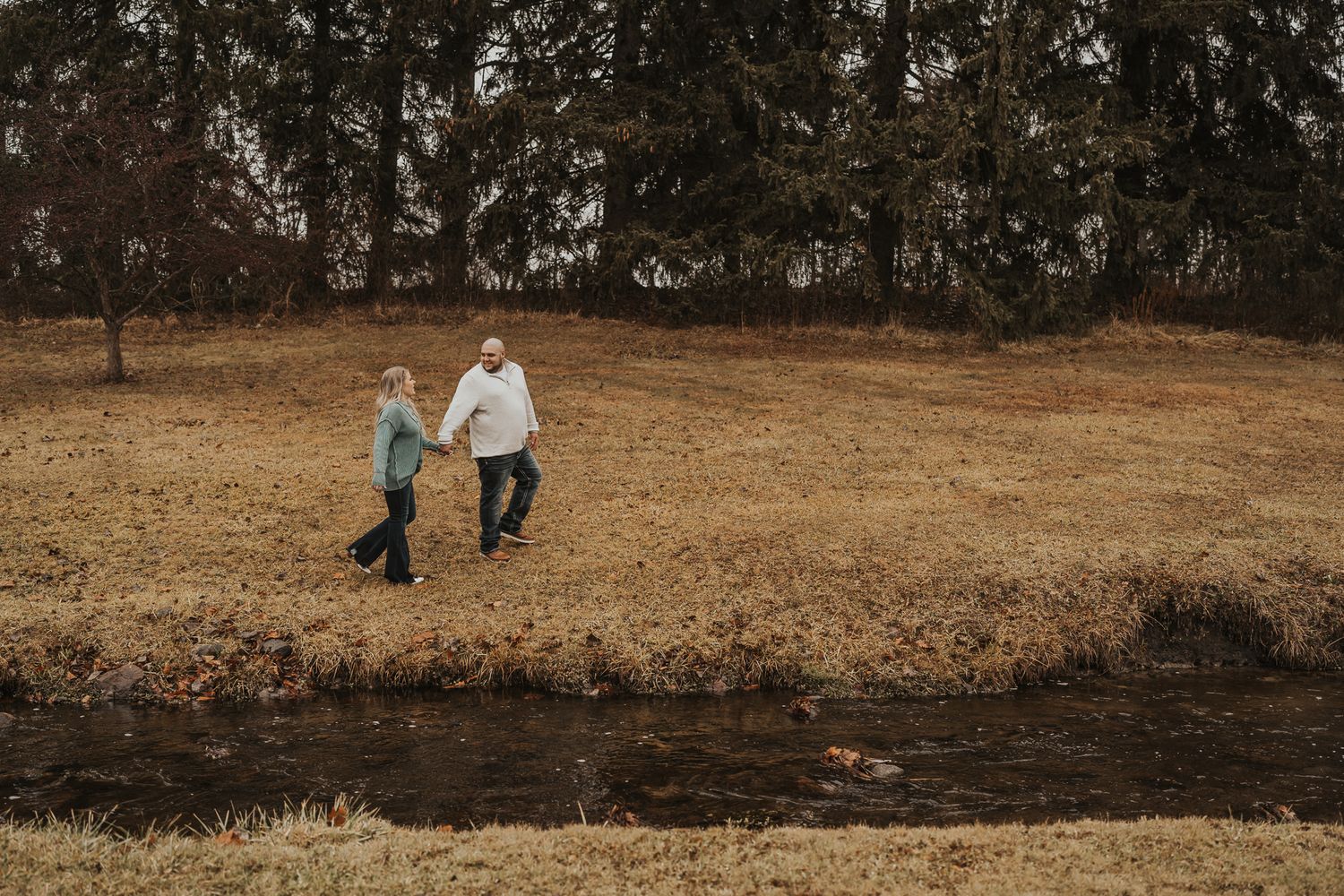 Two people walking along a stream bank on a cloudy autumn day.