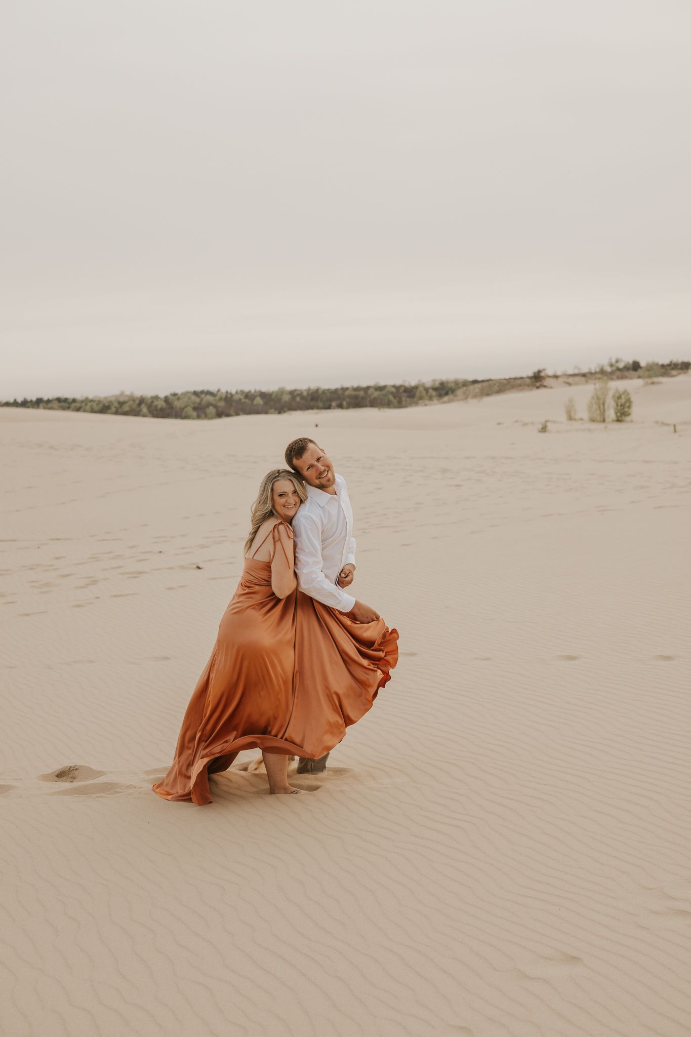 Couple in flowing orange dress and white shirt posing romantically on desert sand dunes.