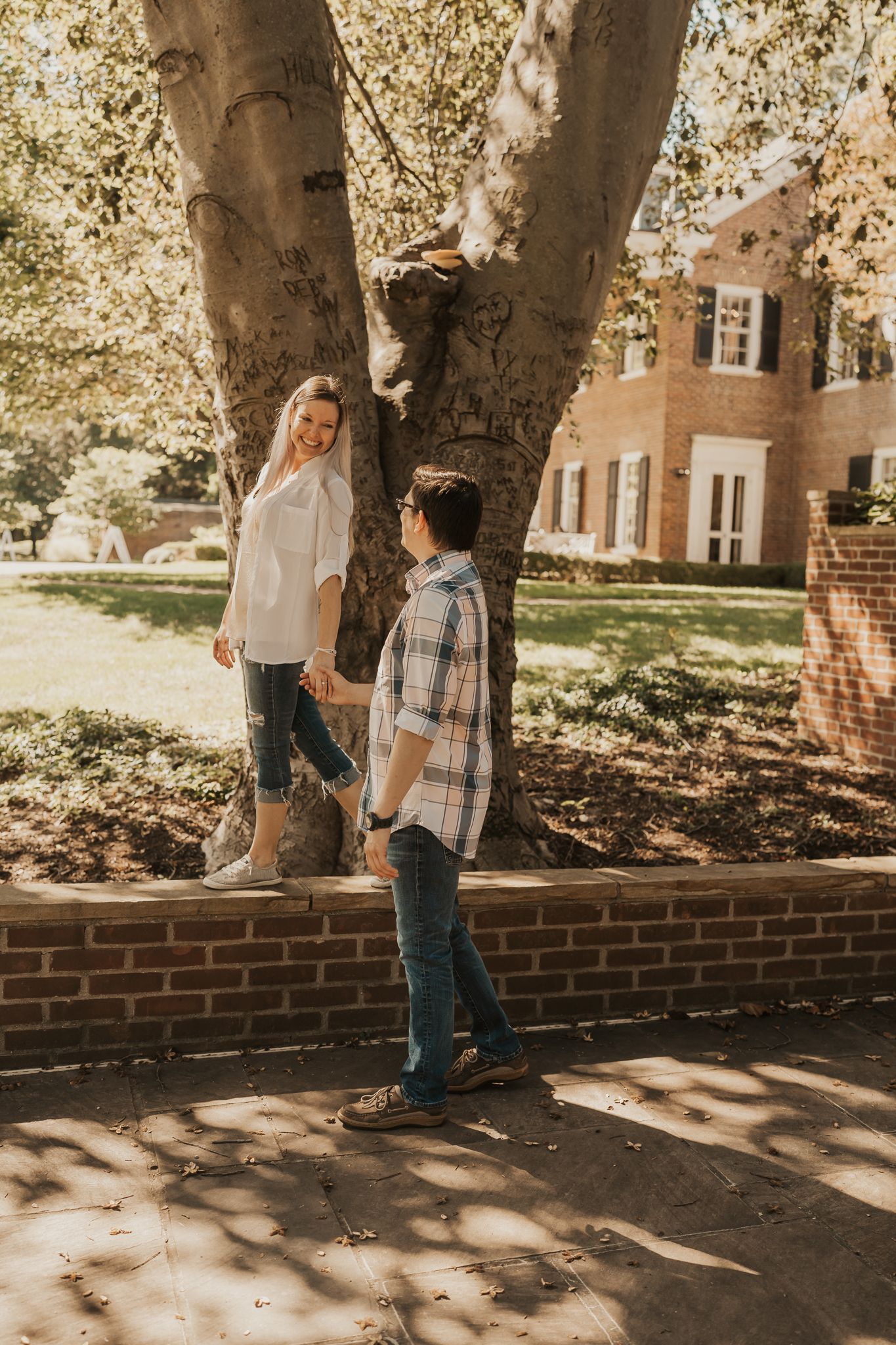 Couple walking along a brick wall beneath large shade trees on a sunny day.