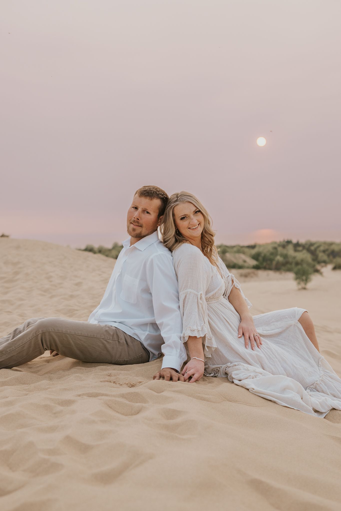 Couple relaxing on sand dunes at sunset with moon visible in pastel sky.