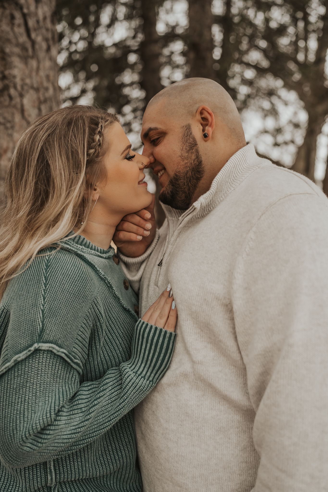 Intimate moment captured between couple in cozy sweaters against forest backdrop.
