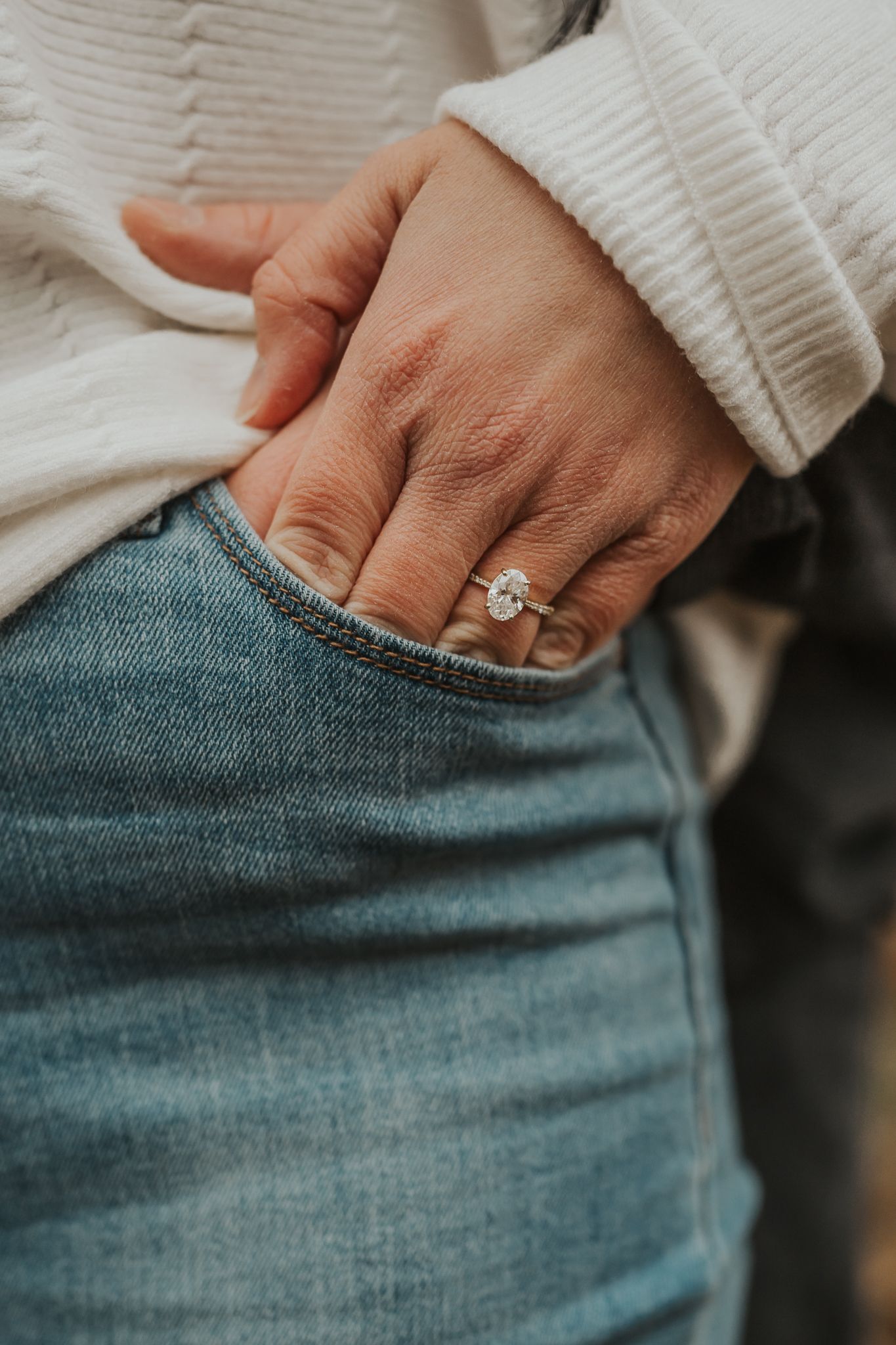 Close-up of a hand wearing an engagement ring against blue jeans and white sweater.