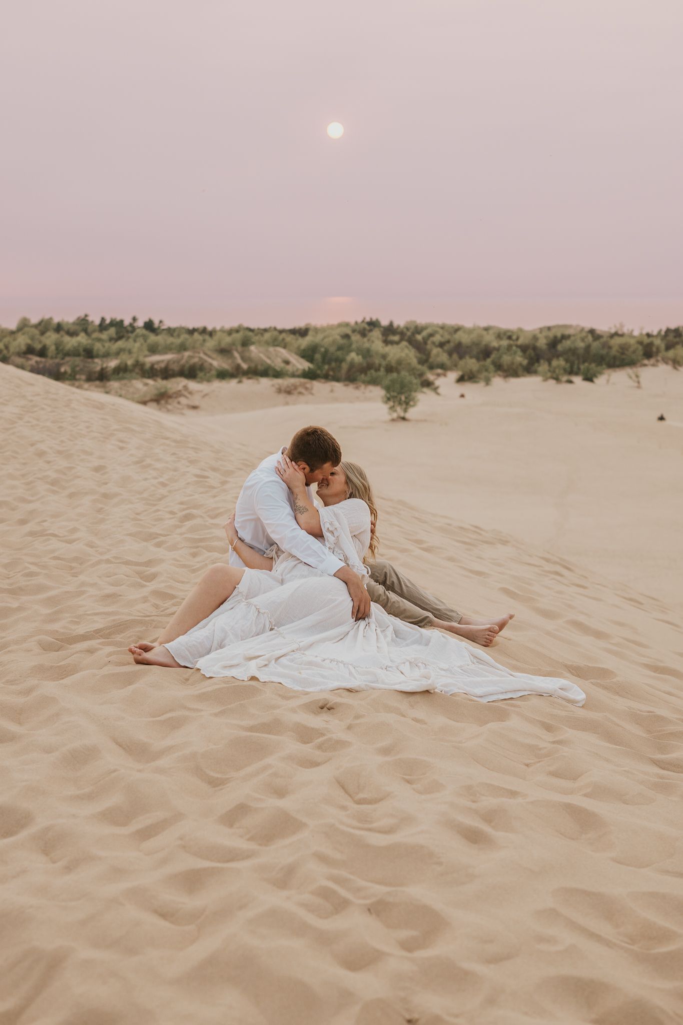 Romantic couple lounging on sandy beach during sunset in light colored outfits.