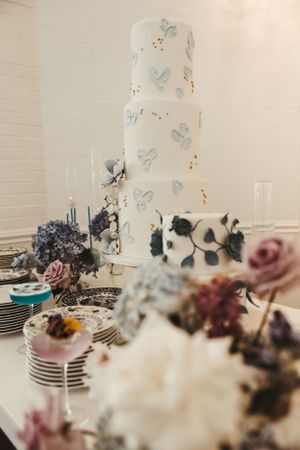 White wedding cake decorated with blue butterflies and surrounded by floral arrangements.