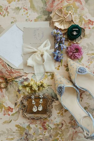 Wedding accessories including flowers, jewelry, and stationery arranged on a floral patterned surface.