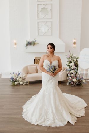 Bride in elegant lace mermaid wedding gown poses in white room with floral arrangements and soft lighting.