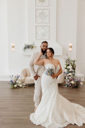 Wedding couple embraces in elegant white venue with hardwood floors and floral arrangements.
