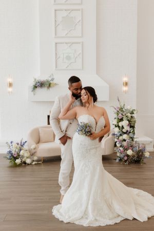 Romantic couple poses together in formal wedding attire in bright white room with floral decor.