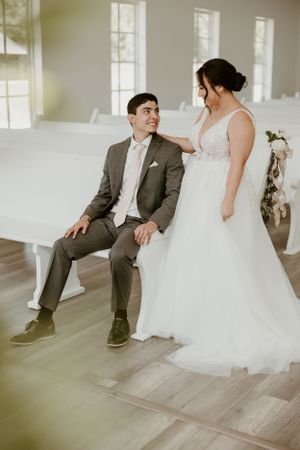 Romantic wedding couple sharing a tender moment on white bench in sunlit venue.