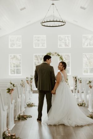 Wedding couple standing in beautiful white chapel with floral arch and chandelier.