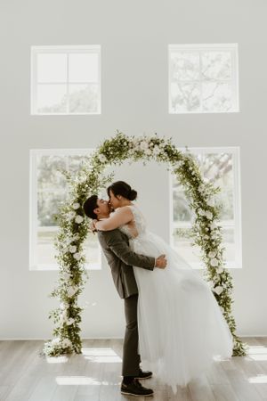 Romantic wedding couple embracing under floral arch with natural light streaming through windows.