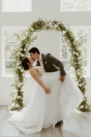 Romantic dip kiss under floral arch in sunlit white chapel.