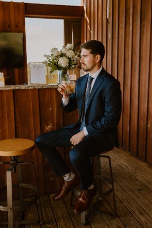 Groom in navy suit sitting on bar stool holding drink in rustic venue.