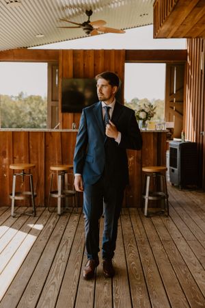 Groom in navy suit adjusting tie while standing on wooden deck.