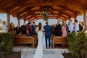 Wedding guests seated in wooden pews under a rustic covered outdoor pavilion.