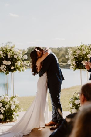 Newlyweds share their first kiss at an outdoor lakeside ceremony surrounded by white floral arrangements.