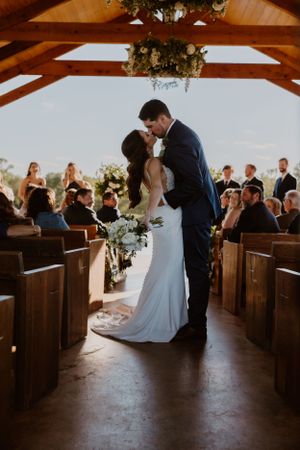 A romantic wedding ceremony kiss in a wooden chapel with guests seated on pews.
