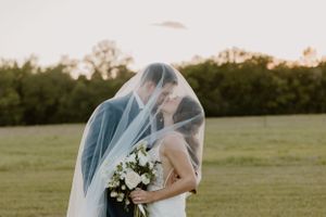 Romantic sunset wedding photo of couple embracing under flowing veil in open field.