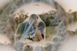 Dreamy double exposure wedding portrait through veil against sunset landscape.